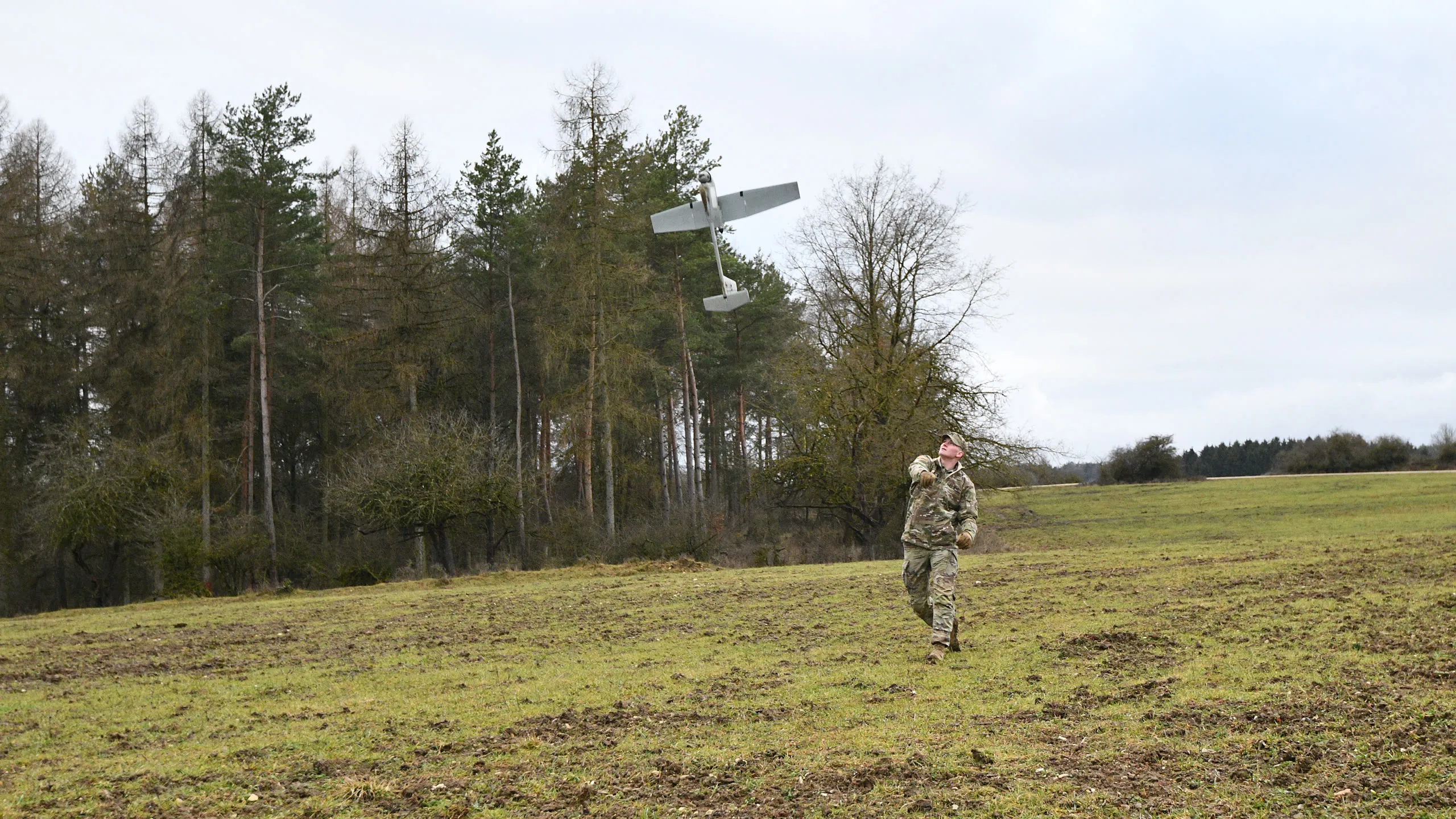 A fixed wing reconnaissance drone released by hand over an open field at the treeline.