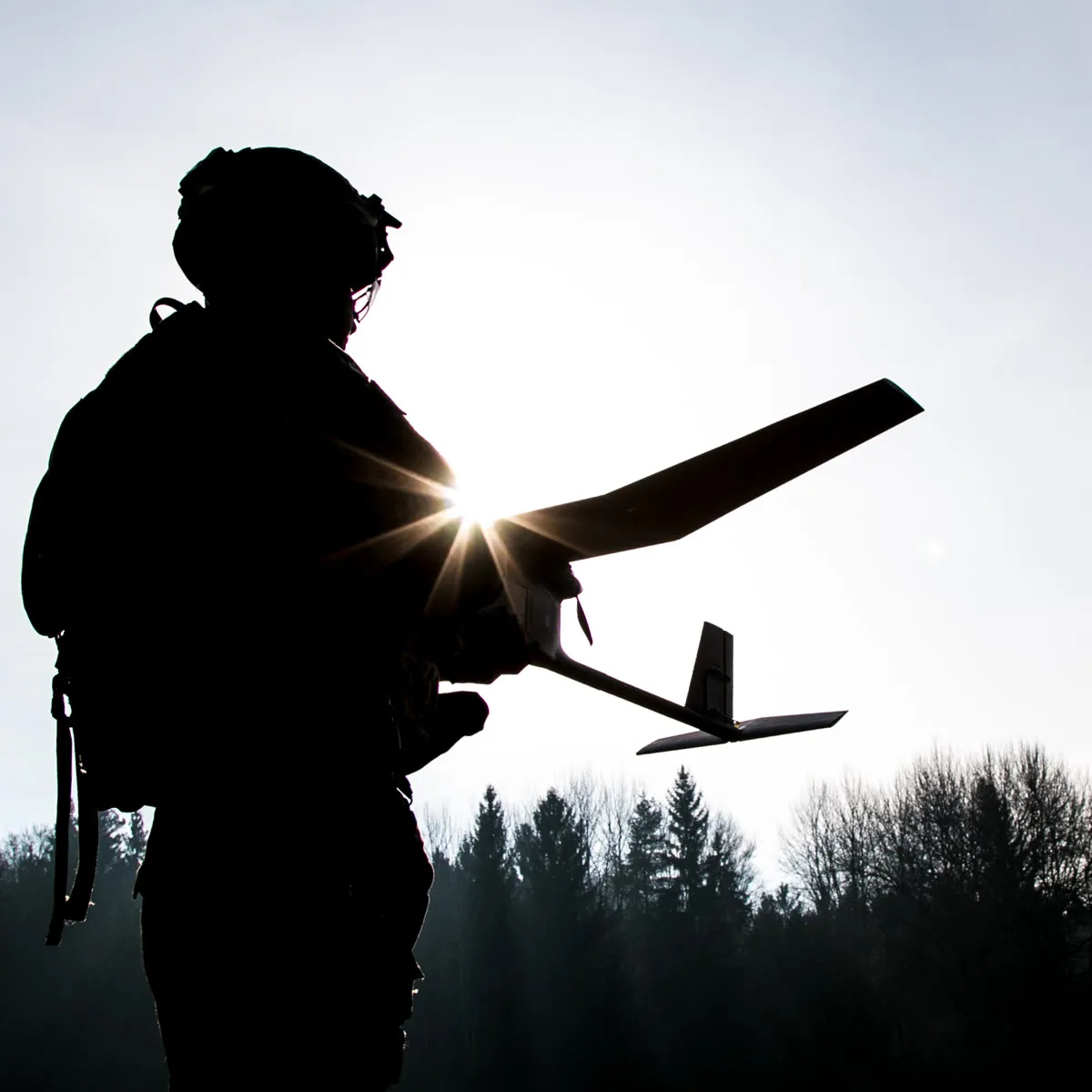A soldier in silhouette against the sun, holding a fixed wing drone.
